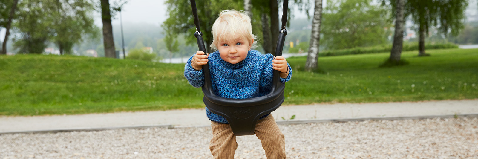 A young toddler plays on a toddler swing cradle, he is looking directly at a camera.